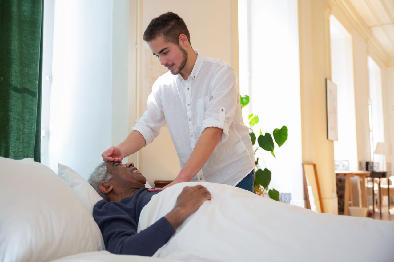our-journey-03 A young man supports an elderly man in bed, offering care in a bright room.